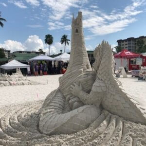 Sand sculpture of a large dragon in front of a tall castle tower.