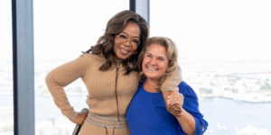 Oprah Winfrey embraces another woman in blue, both smiling in studio with waterfront windows behind